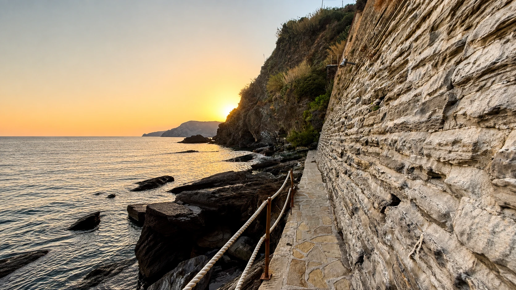 Sentiero costiero tra le rocce al tramonto, con il mare e il sole all'orizzonte